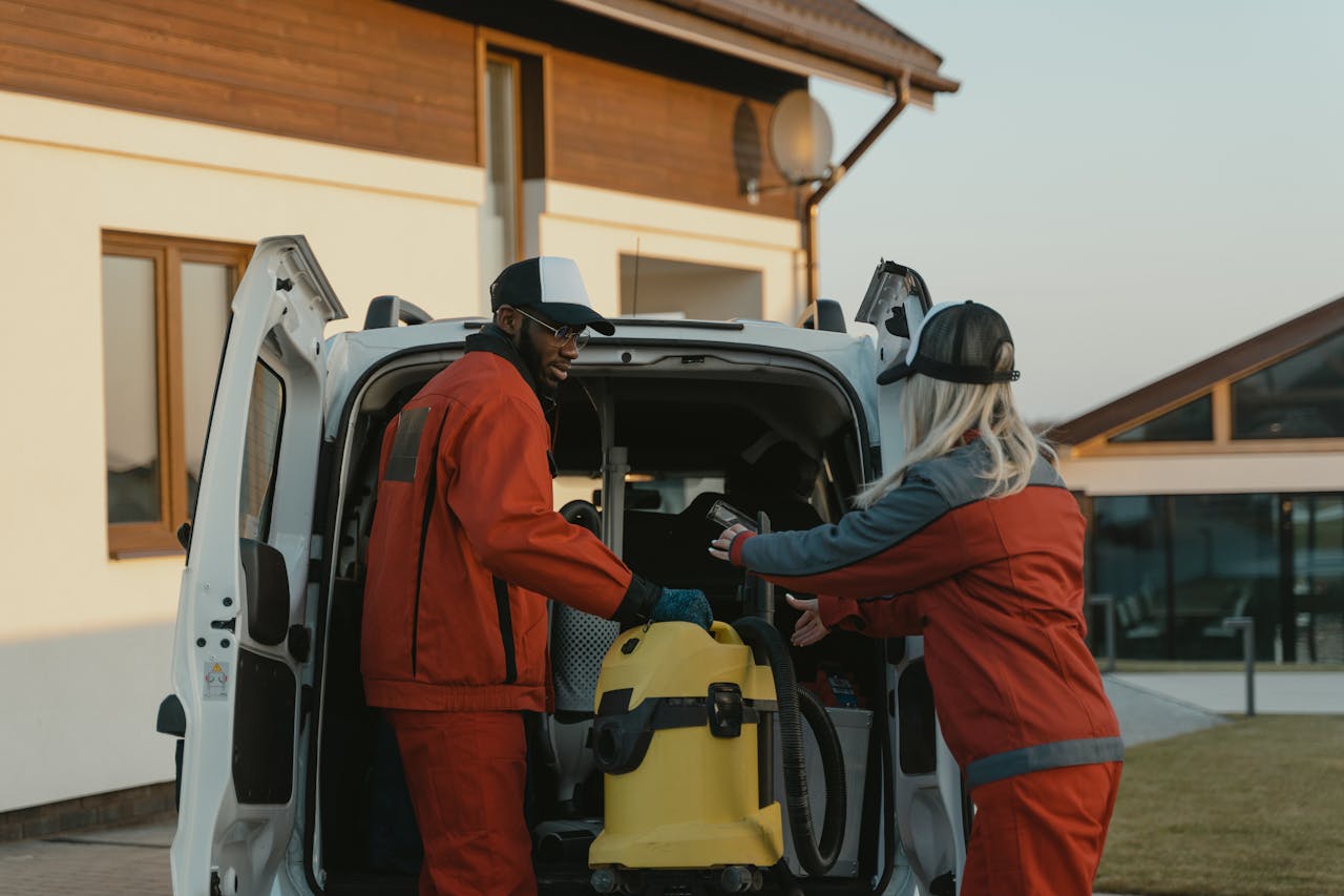 about-02 Two cleaners in red uniforms load a vacuum into a white van outdoors.